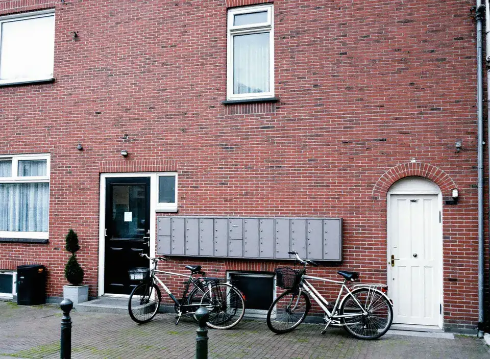 De ingang van een bakstenen portiekflat met grijze brievenbussen en twee fietsen op de stoep