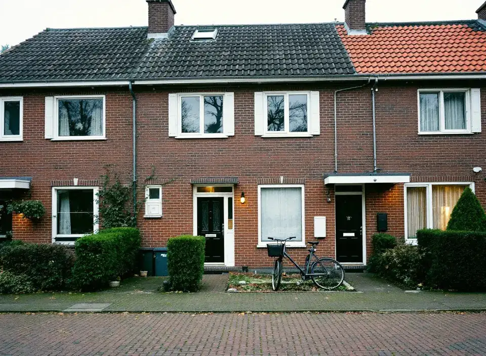 Een rij bakstenen eengezinswoningen uit de jaren zestig met witte kozijnen, heggen en een fiets voor de deur